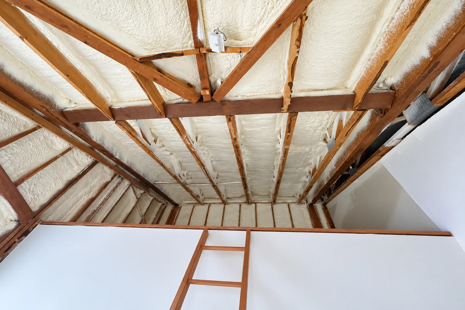 White spray foam fills the roof bays between exposed wooden rafters above a finished room.