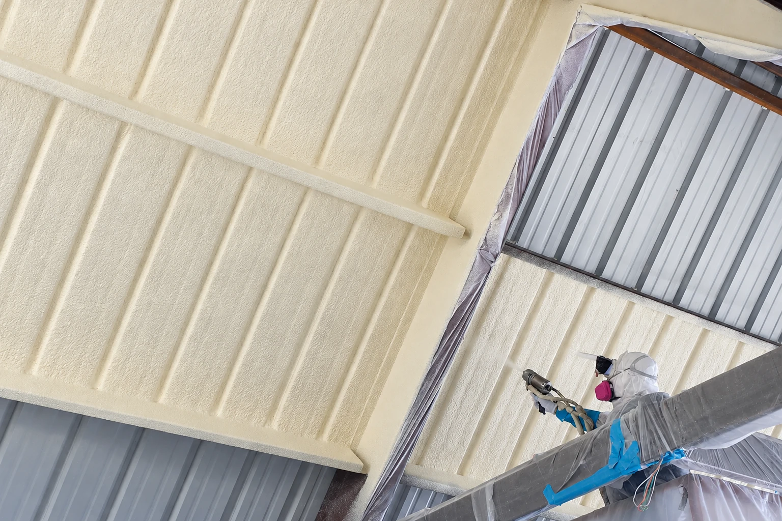 Worker in protective gear sprays cream-colored closed-cell foam onto the underside of a metal roof.