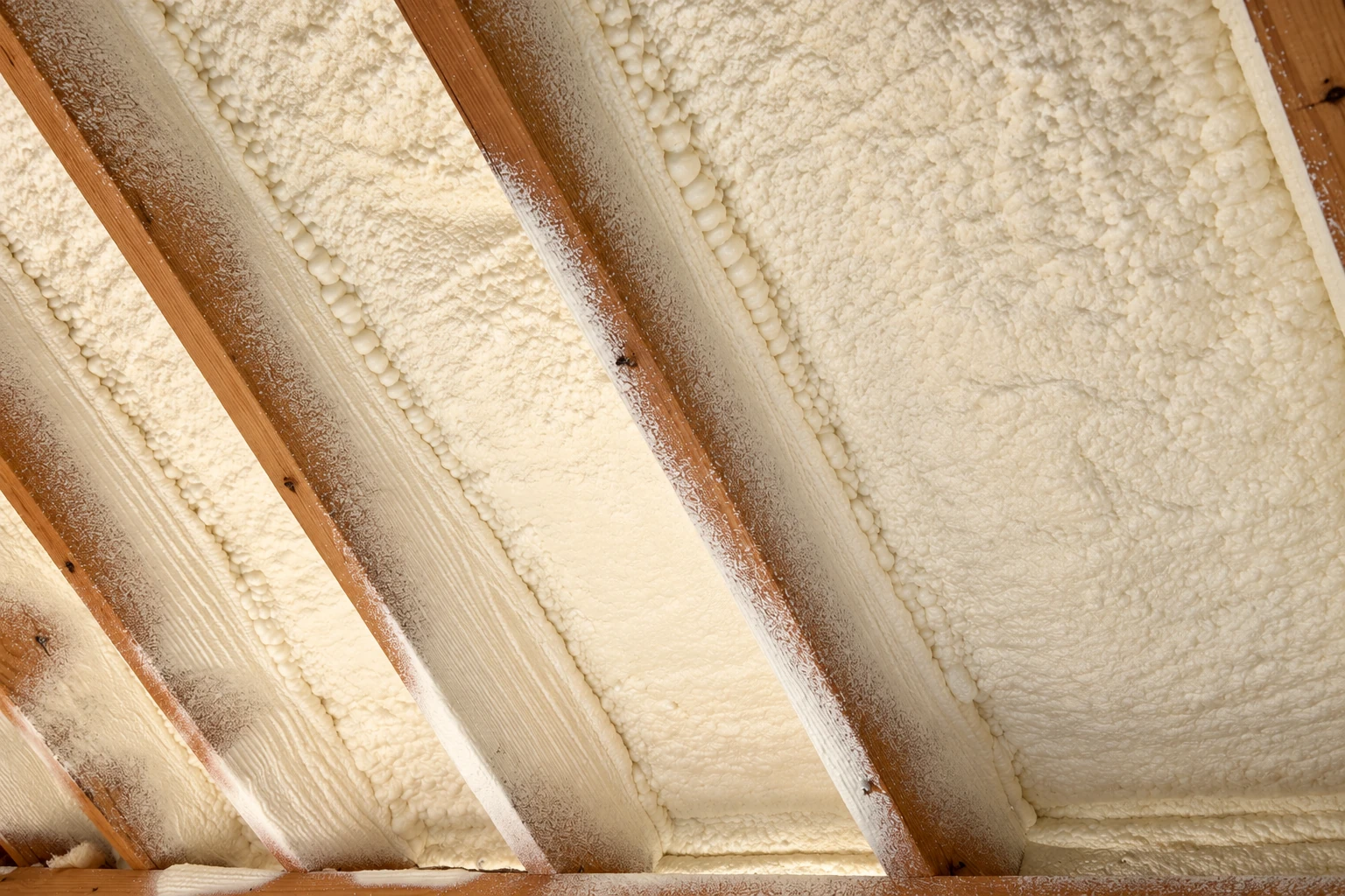 Cream-colored spray foam insulation fills the spaces between exposed wooden rafters.