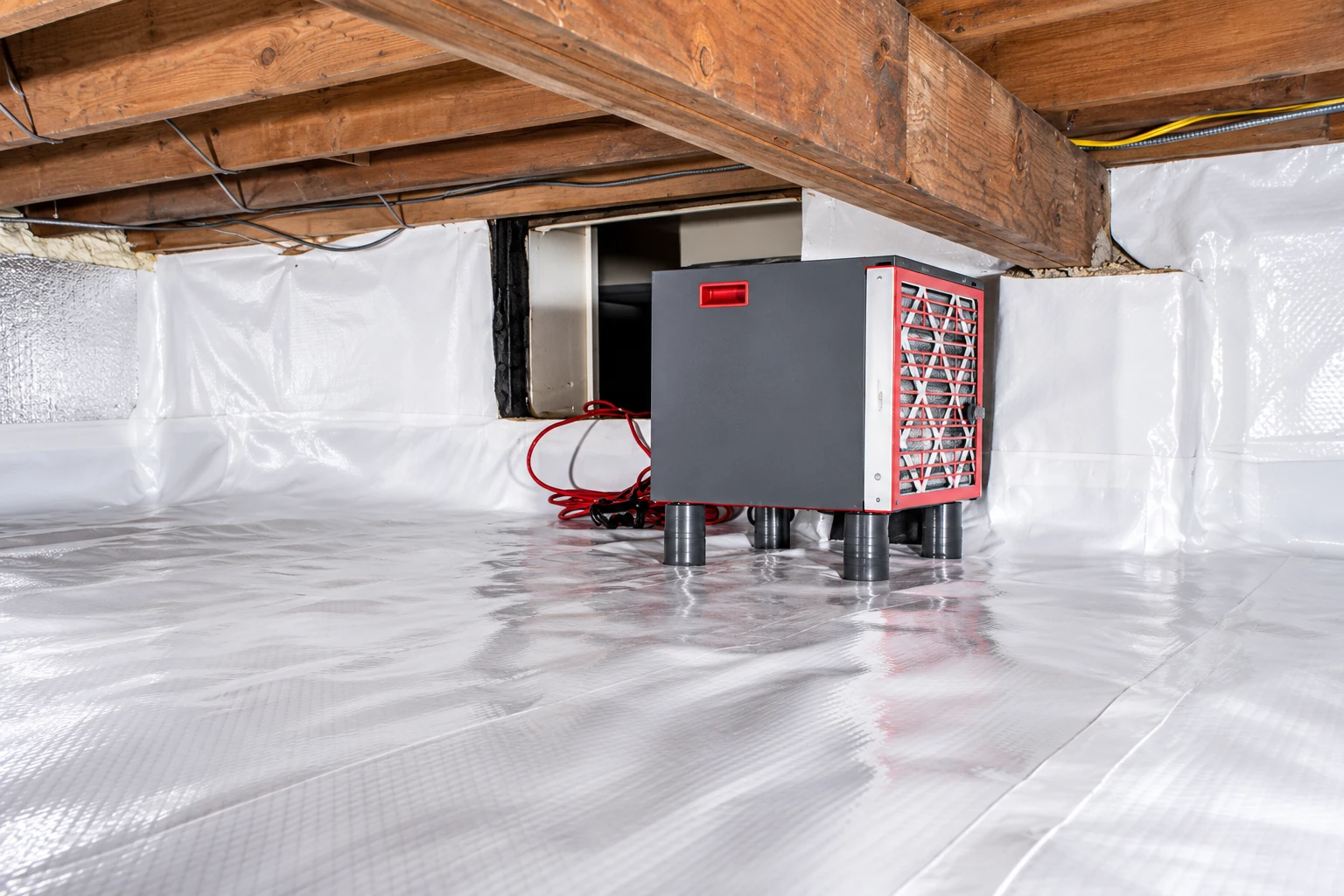 A crawl space floor and walls are covered with reflective vapor barrier beside a gray and red dehumidifier.